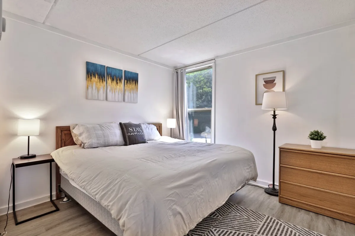 A calm and inviting bedroom featuring a bed with decorative pillows, artwork above the headboard, a nightstand with lamp, and a dresser. Soft natural light filters in, creating a cozy, serene setting for rest and quiet moments.