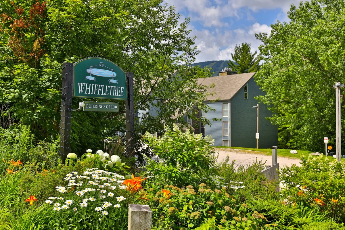 The Whiffletree sign welcomes visitors at the entryway, surrounded by a vibrant flower garden and green foliage. The building facade hints at a friendly, neighborly atmosphere, while the lush landscaping sets a cheerful, inviting tone for arrivals and community events.
