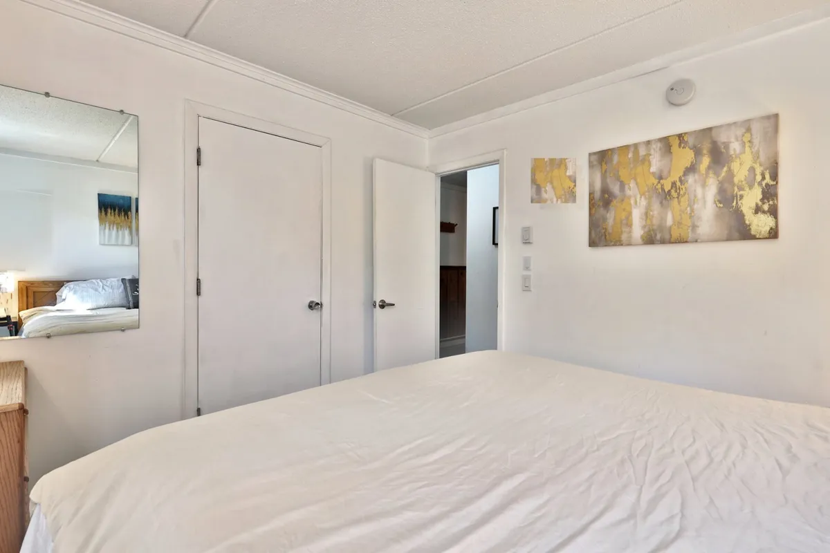 A serene bedroom with a bed dressed in crisp white linens, accentuated by abstract artwork on the wall and a mirror that widens the sense of space. The room feels peaceful and is suited for resting or relaxing, offering a cozy reading nook.