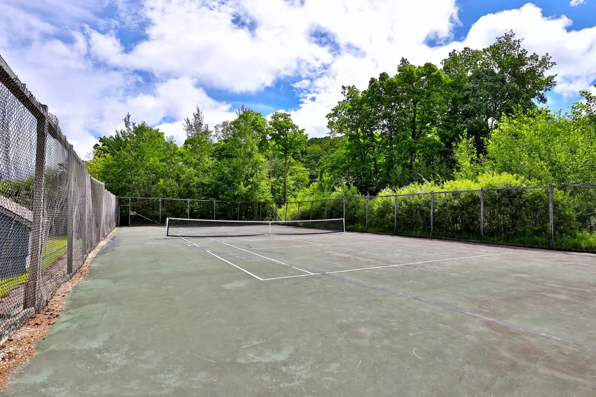 A well-maintained tennis court surrounded by green fencing and lush mature trees, the net stands ready for a friendly match while the peaceful setting encourages both exercise and social gatherings.