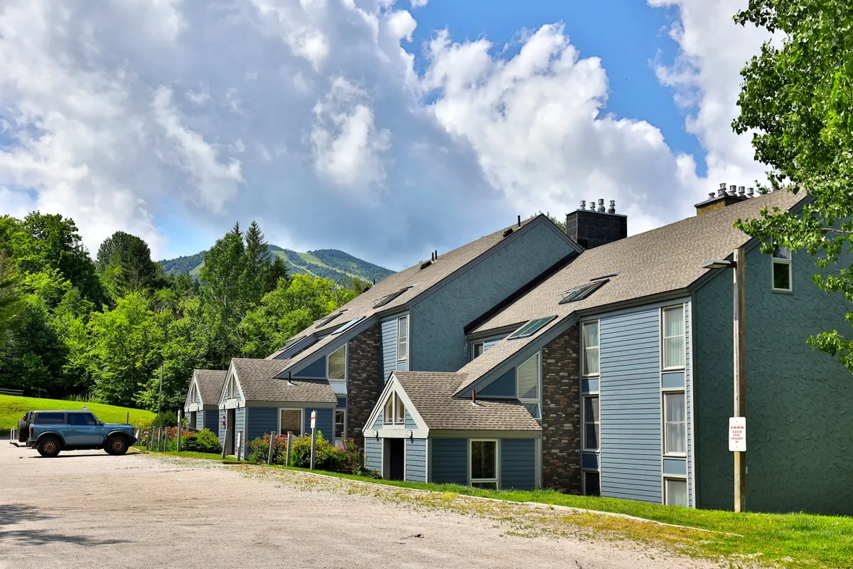 A wide-angle view showcases multi-unit buildings nestled against a dramatic mountain backdrop, surrounded by lush greenery under a cloudy sky. The image conveys a sense of scale, emphasizing the property’s integration with its scenic, mountainous environment—ideal for outdoor adventures.