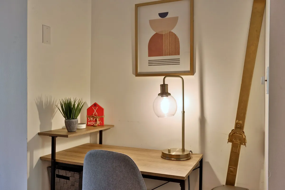 A calm, productive workspace is displayed, featuring a desk with a lamp, modern abstract wall art, a small plant, and a decorative house accessory. The setup is ideal for remote work or moments of inspiration while staying cozy indoors.