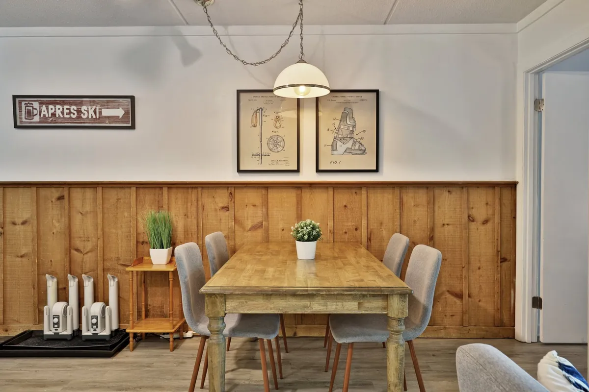 A warm dining area centered around a wooden table, accented by ski-themed artwork and a glowing light fixture. The ambiance is set for sharing meals, playing games, or gathering with friends for memorable moments after an eventful day in Killington.