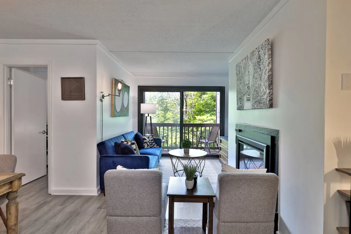 A cozy living area with a blue sofa, large windows offering views of surrounding nature, a coffee table, and a fireplace. The abundant natural light and view of the woods create a welcoming space that seamlessly bridges indoor comfort with the outdoors.