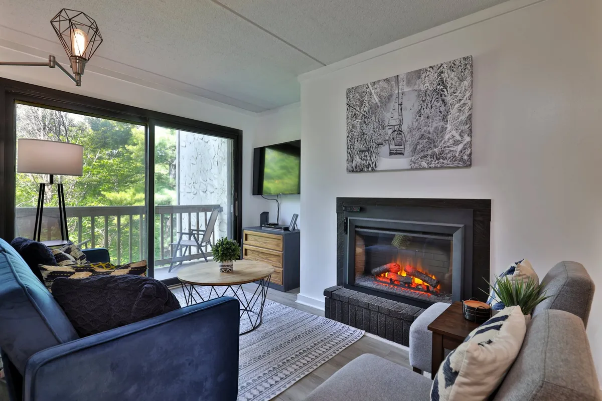 This image features a warm and inviting living room with a fireplace, a comfortable sofa, a coffee table, wall artwork, and a TV setup. Sliding doors in the background hint at access to a balcony with natural views, supporting both relaxation and social activities.