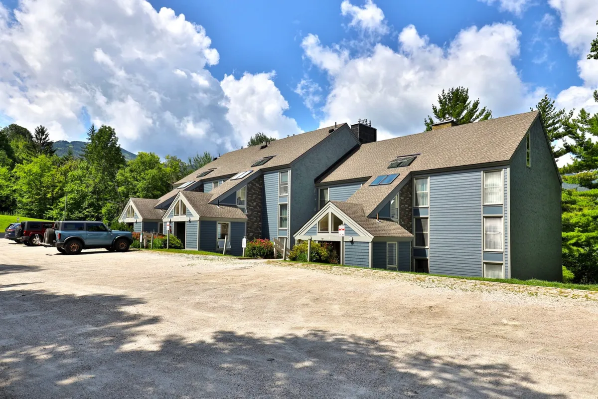 Depicting a multi-story building with a landscaped entrance, parked vehicles, and a welcoming atmosphere, this image places Whiffletree in a lived-in, community context. The scene suggests arrival after a day of skiing or snowboarding, with easy access for families and groups.