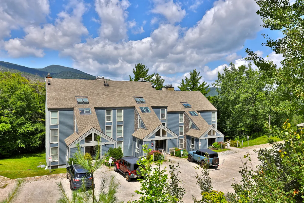 This wide shot features the three-story Whiffletree building set against a tranquil green landscape and mountain backdrop. The parking area, open yard, and surrounding nature suggest ample space for outdoor gatherings, barbecues, and easy access to nearby trails.