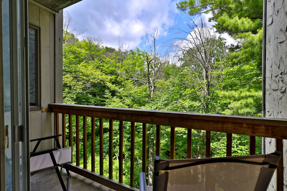 The image features a tranquil balcony with a wooden railing, surrounded by lush greenery and a sky view. The setting invites guests to relax, sip coffee, or soak in the peaceful outdoor atmosphere with glimpses of the natural landscape.