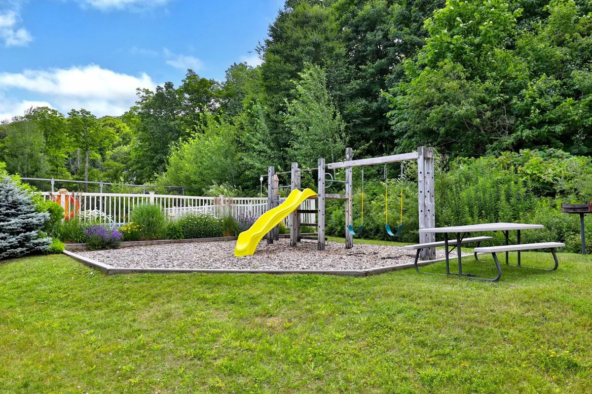 A cheerful playground scene with a bright yellow slide and swing set set on soft grass, surrounded by tall trees. There is a picnic table nearby, ideal for family picnics or supervising children at play, all under a canopy of greenery.