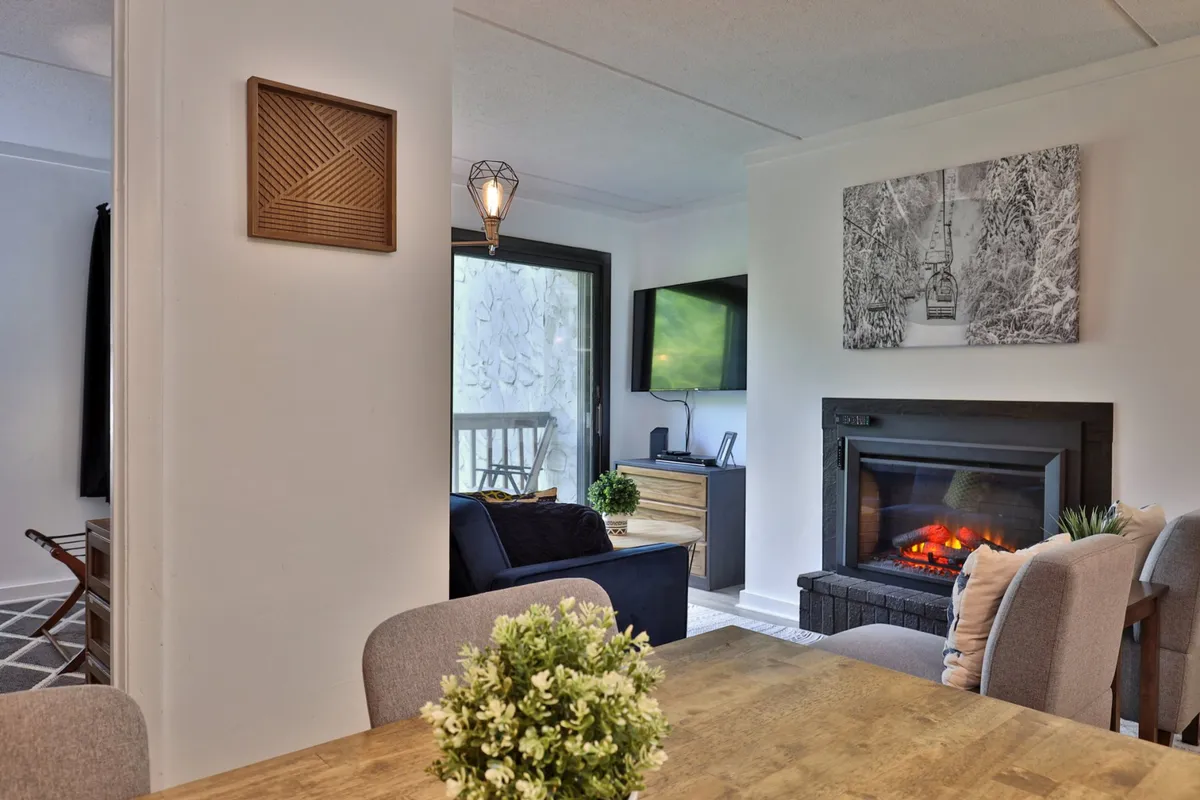 The photo shows a spacious living-dining area with a fireplace, a dining table, a TV, wall artwork, and windows that let in natural light. The furniture is arranged for communal gatherings and comfortable lounging, while the views outside add to the alpine ambiance.