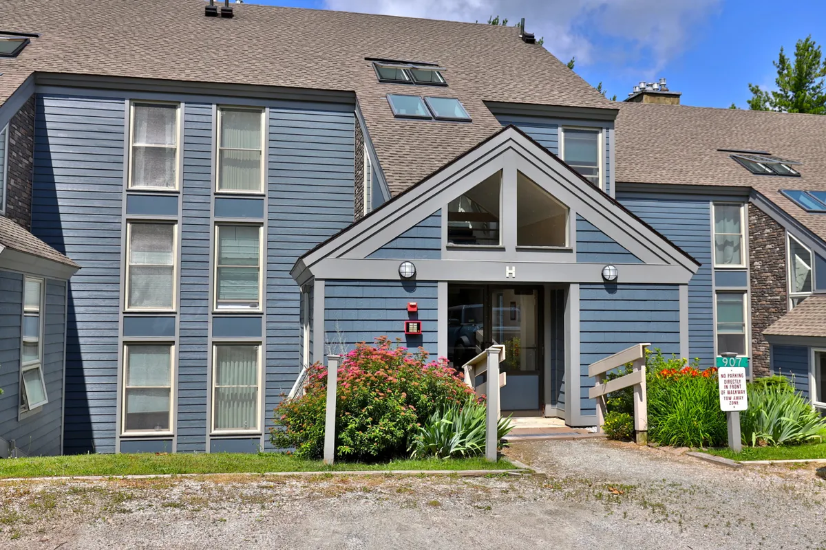A cozy and inviting exterior shot focusing on the entrance to the building, showcasing large windows, colorful flower beds, and architectural features. The setting looks lively yet peaceful, reinforcing the community atmosphere.